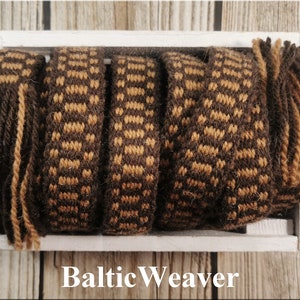 Op de afbeelding: Vijf geweven banden in bruin en beige met franjes aan de uiteinden in een witte houten doos. De doos heeft de tekst "Baltic Weaver" op de voorkant gedrukt.