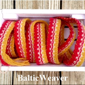 Op de afbeelding: Een close-up van een geweven, veelkleurig lint in een witte doos. Het lint is geel, roze en wit en heeft een geweven patroon. De tekst "Baltic Weaver" is zichtbaar onder de doos.