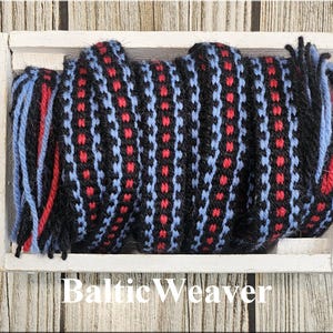 Op de afbeelding: Een geweven sjaal met een geometrisch patroon in zwart, rood en lichtblauw. De sjaal wordt weergegeven in een witte houten bak. De tekst "Baltic Weaver" staat onderaan de afbeelding.