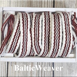 Op de afbeelding: Een rol geweven stof met een herhalend patroon van bordeauxrode en witte verticale strepen, weergegeven in een witte houten kist. De tekst "Baltic Weaver" is zichtbaar onderaan de afbeelding.