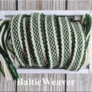 Op de afbeelding: Een geweven textiel in groen en gebroken wit, gepresenteerd in een witte houten bak. Het textiel heeft een herhalend patroon. De tekst "Baltic Weaver" staat in het wit onderaan de afbeelding. Dit textiel wordt waarschijnlijk gebruikt voor knutselen of decoratie.