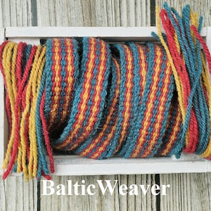 Op de afbeelding: Een close-up van een geweven wandkleed in een witte houten doos. Het wandkleed is gemaakt van rood, geel en blauw garen en heeft een gestreept patroon. De tekst "Baltic Weaver" is zichtbaar onder de doos.