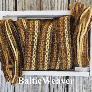 Op de afbeelding: Een geweven wollen band in bruin, geel en wit in een witte houten doos. De band is gemaakt van meerdere samen geweven garenstrengen. De tekst "Baltic Weaver" is afgedrukt op de onderkant van de doos.