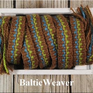 Op de afbeelding: Een close-up van geweven trim in bruin, groen en blauw met franjes. De trim zit in een witte houten doos. De tekst "Baltic Weaver" staat onder de doos.