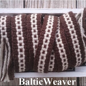 Op de afbeelding: Een close-up van een geweven bies in bruin en wit met franjes. De bies bevindt zich in een houten doos met de tekst "BalticWeaver" op de bodem.