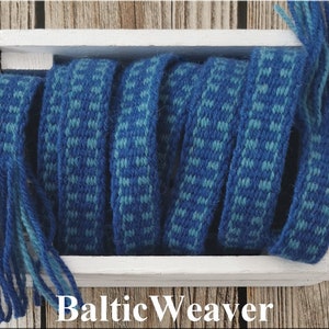 Op de afbeelding: Een close-up van blauw en turquoise geweven stof met franjes aan de uiteinden. De stof is opgerold in een witte houten doos. De tekst "Baltic Weaver" is onder de doos gedrukt.