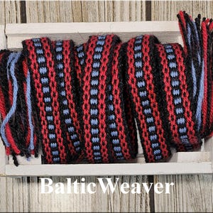 Op de afbeelding: Geweven textiel in een witte bak. Het textiel heeft een herhalend patroon van rood, zwart en lichtblauw. De uiteinden van het textiel hebben losse draden in rood, zwart en lichtblauw. De tekst "Baltic Weaver" staat onderaan.
