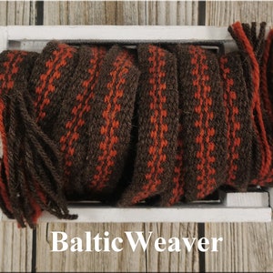 Op de afbeelding: Een close-up van een geweven bruine en oranje stof met kwastjes. De stof zit in een witte houten doos. De tekst "Baltic Weaver" staat onder de doos.