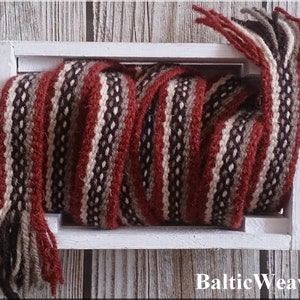 Op de afbeelding: Een geweven wollen band in de kleuren rood, bruin, wit en zwart met franjes aan beide uiteinden. De band is opgerold en tentoongesteld in een witte houten doos. De tekst "BalticWeaver" is onderaan de afbeelding gedrukt.