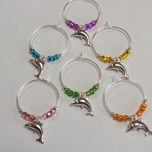 May include: Six silver wine glass charms with a dolphin charm and colorful beads. The charms are in the colors purple, blue, yellow, green, pink, and orange.