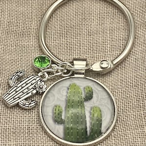 May include: A silver key chain with a round charm featuring a green cactus illustration. The charm is attached to a silver ring with a green crystal charm.