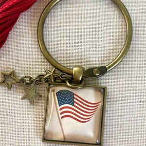 May include: A bronze-toned metal keychain featuring a circular ring and a square pendant. The pendant displays a graphic of the American flag on a light background. Two small bronze star charms are attached to the ring near the pendant.