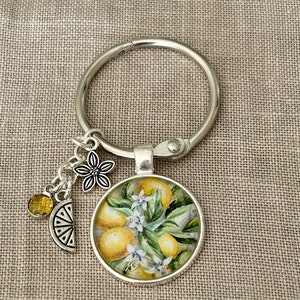 May include: A silver key chain with a round charm featuring a painting of lemons and blossoms. The charm is attached to a silver ring with a small silver flower charm and a yellow glass bead.