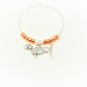 May include: A silver dachshund charm hangs from a copper and white beaded wine glass charm.
