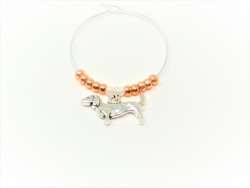 May include: A silver dachshund charm hangs from a copper and white beaded wine glass charm.