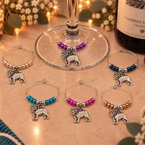 May include: A set of six circular wine glass charms, each featuring a silver-toned metal pug dog charm suspended from a wire hoop. The hoops are decorated with small, round beads in various colors, including metallic silver, purple, dark blue, teal, pink, and bronze.