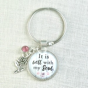 May include: A silver-toned metal keychain featuring a circular pendant with the text It is well with my soul printed over a white wood-grain background. The keychain includes a small pink rhinestone charm and a silver rose-shaped metal charm.