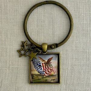 May include: A bronze-toned keychain featuring a square pendant with an American flag design and a bronze-colored ring. The pendant shows a waving flag and an eagle. Three bronze stars dangle from the ring.