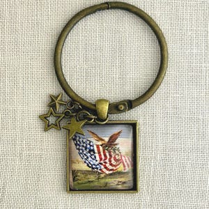 May include: A bronze-toned circular keychain featuring a square pendant with a glass-covered image of a bald eagle clutching an American flag. The keychain includes three small bronze star charms attached to the side of the square frame.