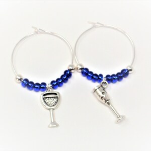 May include: A pair of silver-toned hoop earrings featuring a row of small, metallic blue beads along the bottom curve. Each hoop is adorned with a dangling silver-toned charm, one shaped like a wine glass and the other like a champagne flute.