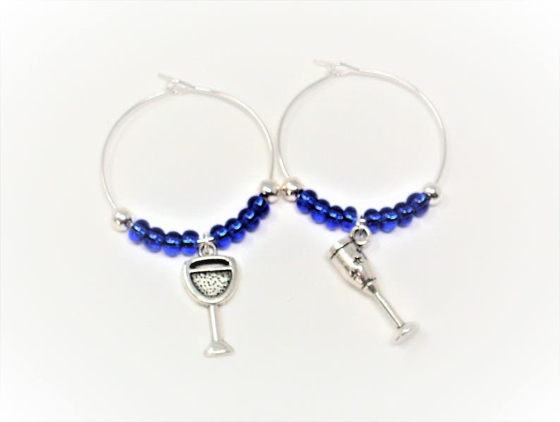 May include: A pair of silver-toned hoop earrings featuring a row of small, metallic blue beads along the bottom curve. Each hoop is adorned with a dangling silver-toned charm, one shaped like a wine glass and the other like a champagne flute.