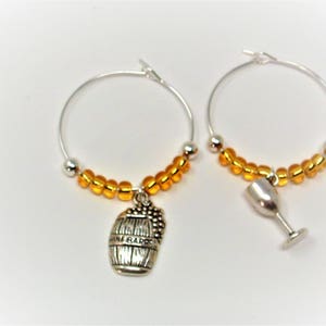 May include: A pair of silver-toned hoop earrings featuring a row of small, translucent amber-colored beads. One hoop includes a silver-toned charm shaped like a wine barrel, while the other hoop features a silver-toned wine glass charm.