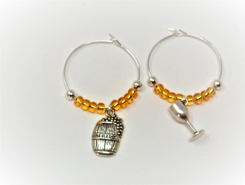 May include: A pair of silver-toned hoop earrings featuring a row of small, translucent amber-colored beads. One hoop includes a silver-toned charm shaped like a wine barrel, while the other hoop features a silver-toned wine glass charm.