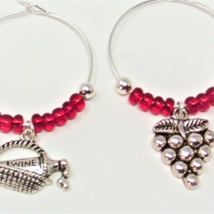 May include: Two silver-toned hoop wine glass charms featuring red glass beads and silver metallic accents. One charm displays a basket with a bottle labeled WINE, while the other charm features a cluster of grapes. The charms are circular in shape.