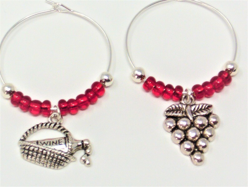 May include: Two silver-toned hoop wine glass charms featuring red glass beads and silver metallic accents. One charm displays a basket with a bottle labeled WINE, while the other charm features a cluster of grapes. The charms are circular in shape.