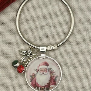May include: A silver-toned metal keychain featuring a circular pendant with a vintage-style illustration of Santa Claus in a red suit. The keychain includes a small silver holly leaf charm, a green crystal bead, and a red crystal bead attached.