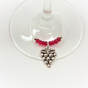 May include: A wine glass charm featuring a silver-toned metal grape cluster pendant suspended from a circular wire hoop. The hoop is decorated with a series of small, round, translucent red beads arranged symmetrically on either side of the charm.