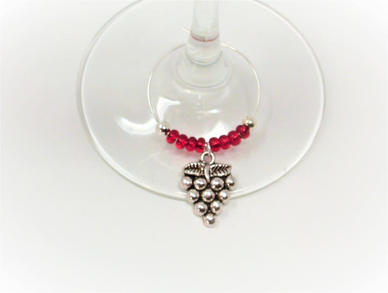 May include: A wine glass charm featuring a silver-toned metal grape cluster pendant suspended from a circular wire hoop. The hoop is decorated with a series of small, round, translucent red beads arranged symmetrically on either side of the charm.