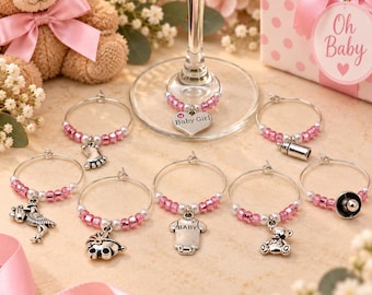 May include: A set of silver-toned metal wine glass charms featuring baby-themed pendants including a heart with the text Baby Girl, a footprint, a baby bottle, a stork, a carriage, a onesie with the text Baby, a teddy bear, and a pacifier.