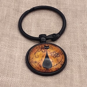 May include: Black metal keychain with a round glass charm featuring a black silhouette of a cat on a vintage floral background.