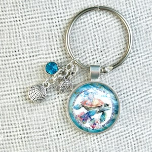 May include: A silver key chain with a round glass charm featuring a watercolor painting of a sea turtle swimming with bubbles. The charm is attached to a silver ring with a small silver seashell and a blue crystal charm.