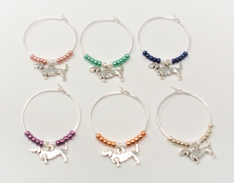 May include: Six silver wine glass charms with dachshund charms. Each charm has a different color of beads: pink, green, blue, purple, orange, and gold.