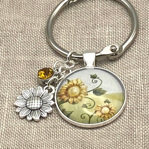 May include: A silver key chain with a round charm featuring a sunflower design. The charm has a clear glass cover and a silver border. The key chain also has a small silver sunflower charm and a yellow crystal charm.