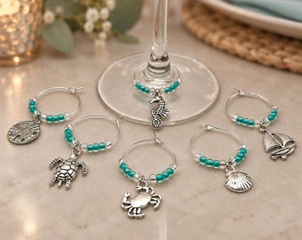 May include: A set of five silver-toned metal wine glass charms featuring nautical and sea-life motifs including a seahorse, turtle, crab, seashell, and sailboat. Each circular charm is accented with small, translucent teal and clear glass beads.