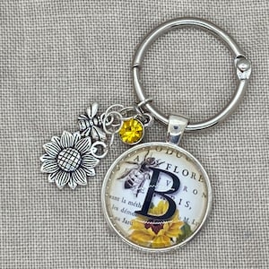 May include: A silver key chain with a charm featuring a bee and sunflower design. The charm has a glass cabochon with a vintage book page background and the letter "B" in black. The text on the page reads "A FLORE V RON B I S, M. L. avant la méth les démon au Jaru".