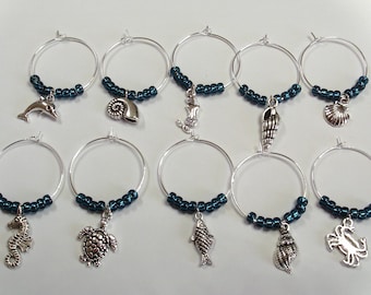 May include: A set of ten silver-toned metal wine glass charms, each featuring a circular hoop adorned with dark teal beads and a unique ocean-themed charm, including a dolphin, various seashells, a mermaid, a seahorse, a turtle, a fish, and a crab.