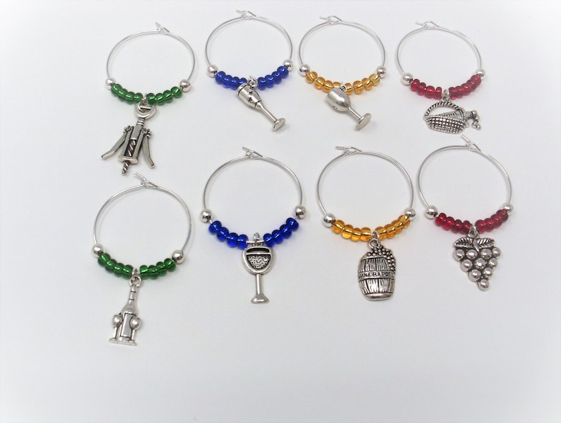 May include: A set of eight circular metal wine glass charms featuring silver-toned charms shaped like wine bottles, glasses, barrels, grapes, and corkscrews. Each charm is accented with a row of small, colorful glass beads in green, blue, orange, or red.