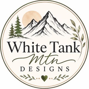 May include: A circular logo featuring a black and white mountain range, two pine trees, and a pale orange sun. The text reads White Tank Mtn DESIGNS in black lettering. A green heart and leafy branches decorate the bottom of the circular frame.