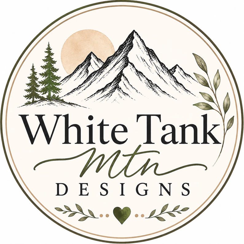 May include: A circular logo featuring a black and white mountain range, two pine trees, and a pale orange sun. The text reads White Tank Mtn DESIGNS in black lettering. A green heart and leafy branches decorate the bottom of the circular frame.