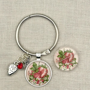 May include: A silver-toned metal keyring featuring a circular pendant with a strawberry illustration, accompanied by a small silver strawberry charm and a red crystal bead. A matching circular strawberry-themed pin with a needle is positioned nearby.