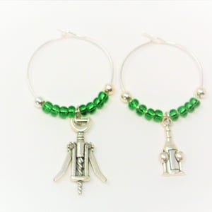 May include: A pair of silver-toned hoop wine charms featuring green glass beads. One charm displays a metal corkscrew pendant, while the other charm features a metal wine bottle pendant. These circular wire hoops are designed for drink identification.