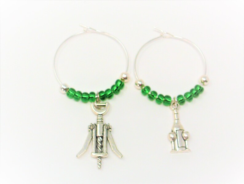 May include: A pair of silver-toned hoop wine charms featuring green glass beads. One charm displays a metal corkscrew pendant, while the other charm features a metal wine bottle pendant. These circular wire hoops are designed for drink identification.
