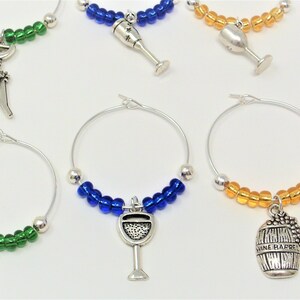 May include: A collection of silver-toned metal wine glass charms featuring circular wire hoops adorned with colorful glass beads. Each charm includes a unique dangling pendant shaped like wine-related items such as glasses, barrels, and corkscrews.