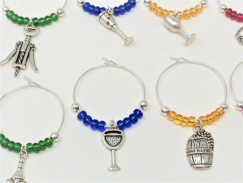 May include: A collection of silver-toned metal wine glass charms featuring circular wire hoops adorned with colorful glass beads. Each charm includes a unique dangling pendant shaped like wine-related items such as glasses, barrels, and corkscrews.