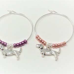 May include: Two silver wine glass charms with dachshund charms. One charm has a purple and white beaded chain, the other has a copper and white beaded chain.