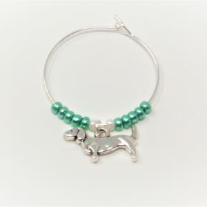 May include: A silver charm of a dachshund dog hanging from a silver wire hoop with green and white beads.